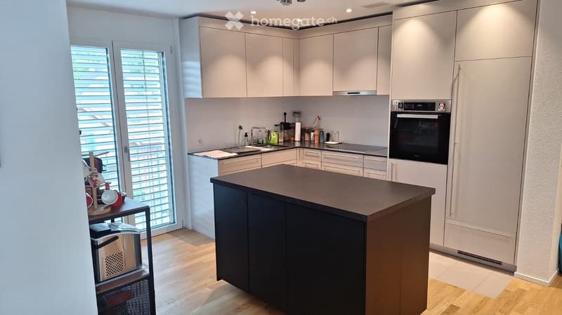 Modern kitchen with white cabinets, a central island, and various appliances including an oven and refrigerator. The space features a sink with a faucet, and several bottles and cups are placed on the counter.