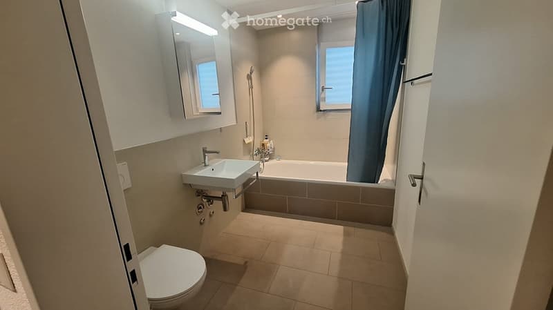 Modern bathroom with a toilet, sink, bathtub, and mirror. Features tiled walls and floor, a window with a blue curtain, and a showerhead above the bathtub.