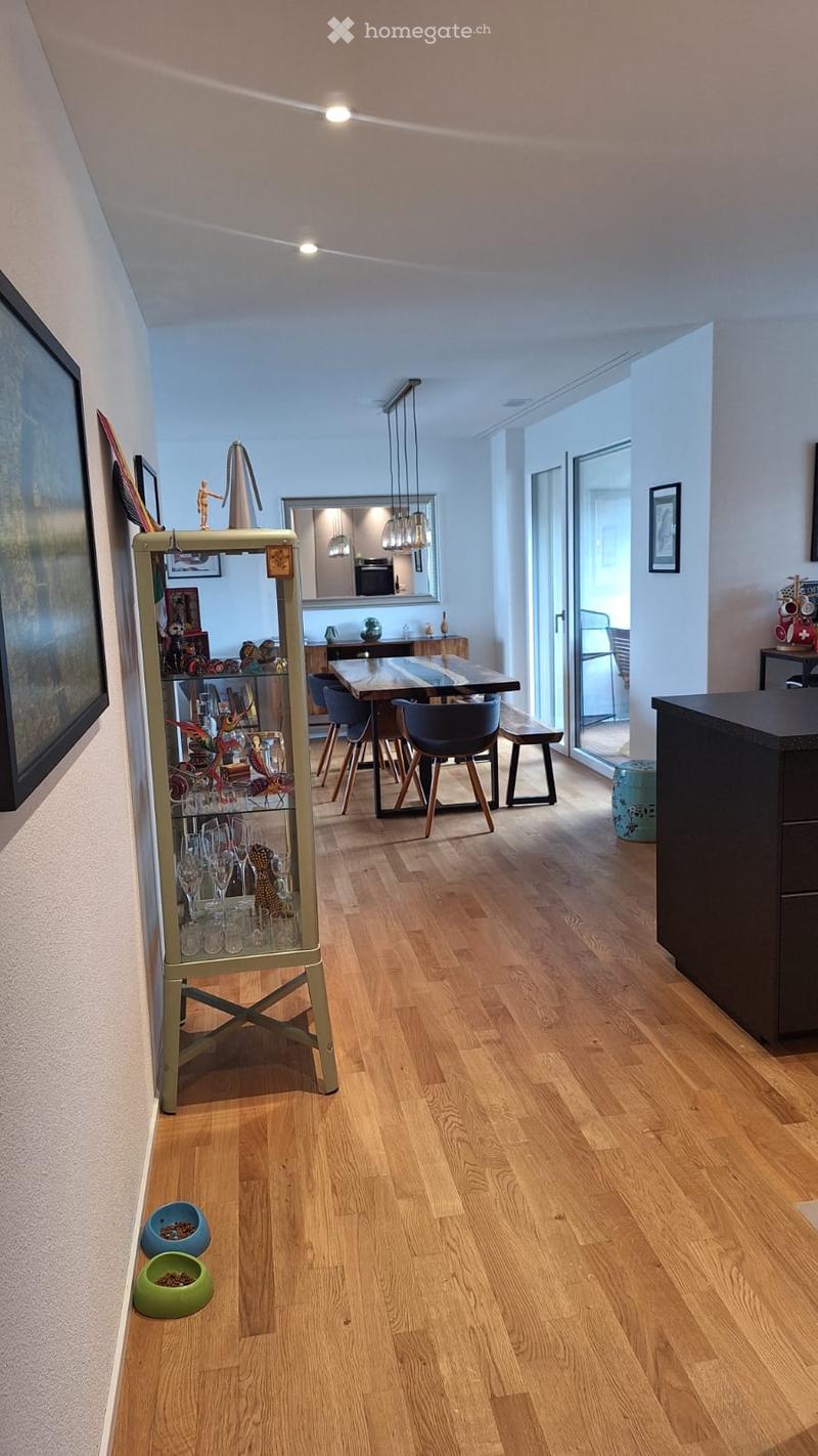 Bright hallway with wooden flooring, leading to a dining area with a table and chairs, and a display cabinet with glassware and decorative items. A pet food bowl is placed on the floor near the entrance.