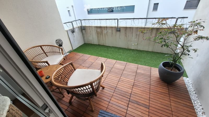 Small wooden deck with two wicker chairs, a small table, and a potted plant, surrounded by a low green artificial grass area and a white building wall.