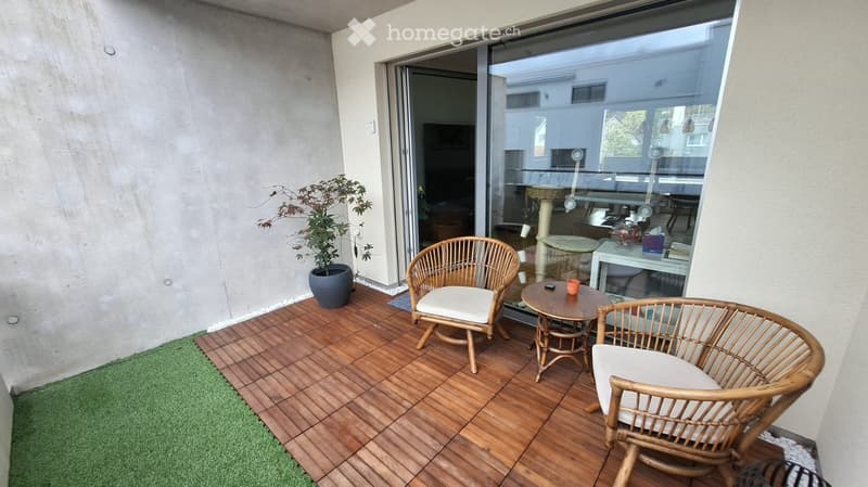 Balcony with wooden decking, two wicker chairs with white cushions, small round table, potted plant, and a view into the adjacent room.
