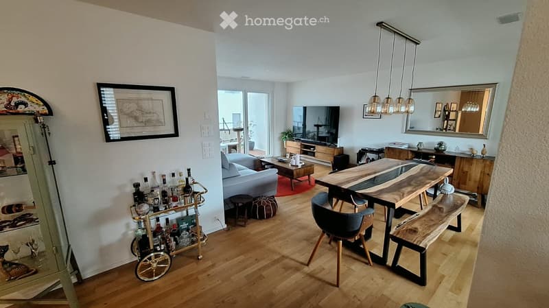 Modern living room with hardwood floors, a grey sofa, coffee table, ottoman, a large wooden dining table with chairs and bench, a bar cart, and a TV area with a cabinet and mirror.