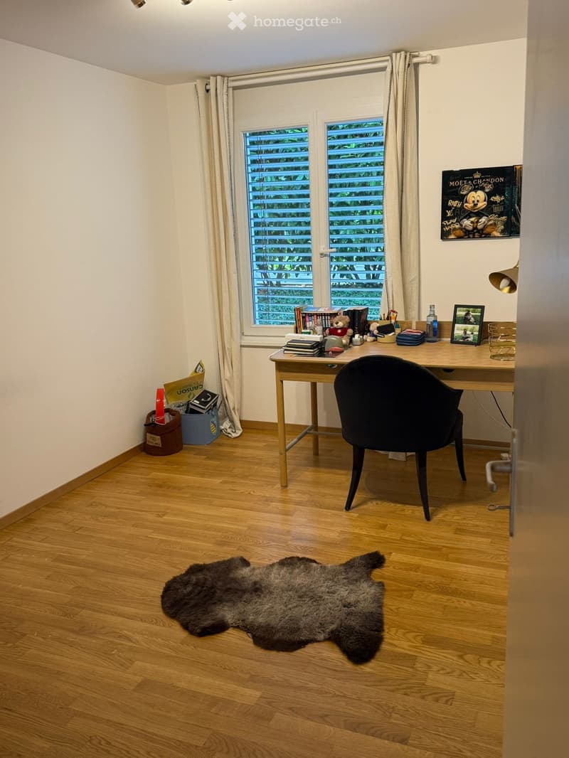 A well-lit study or home office with wooden flooring, a large window with blinds and curtains, a wooden desk with a black chair, books, a lamp, and a fur rug on the floor.