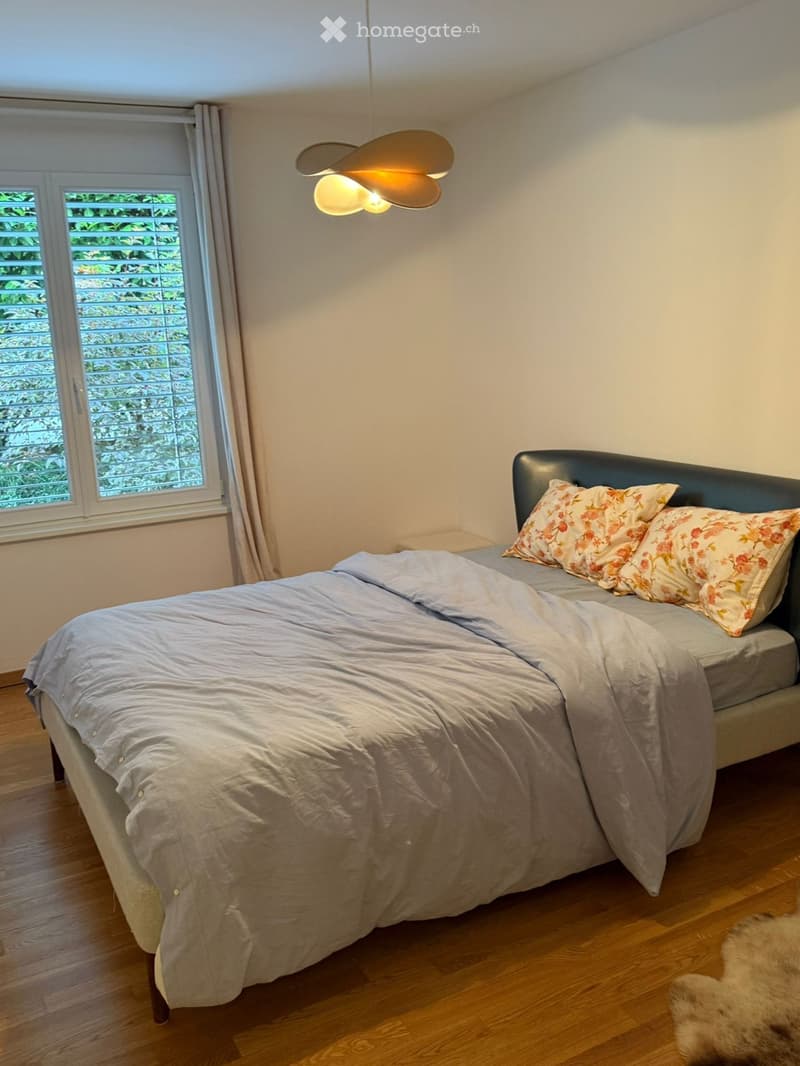 A well-lit bedroom with a double bed, light grey duvet, floral pillows, wooden flooring, and a large window with blinds and a curtain. A modern pendant light hangs from the ceiling.