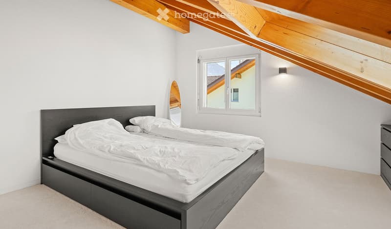 Modern bedroom with a large bed, white bedding, a mirror, and a nightstand. The room features a slanted wooden ceiling and large windows with a scenic view.