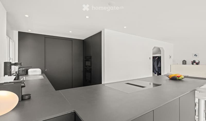 Modern kitchen with grey countertops, white walls, black cabinetry, and built-in appliances. Features a central island with a sink and a small seating area with stools.