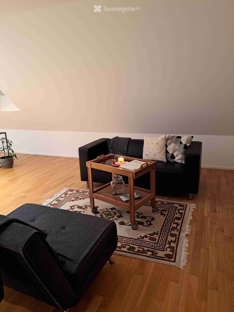 Modern living room with wooden flooring, a dark grey sofa, an ottoman, a coffee table with a candle and jar, and a patterned rug.