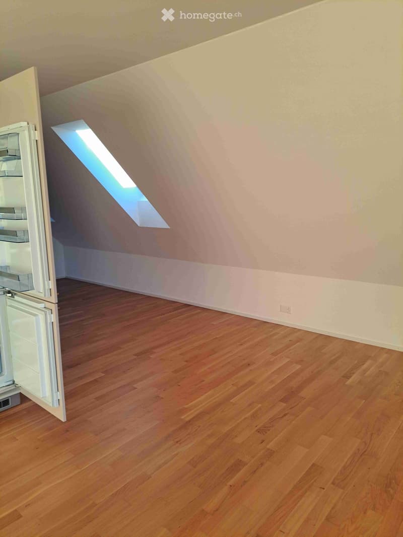 Empty room with wooden flooring, slanted ceiling, and a skylight window. A refrigerator is partially visible on the left side.