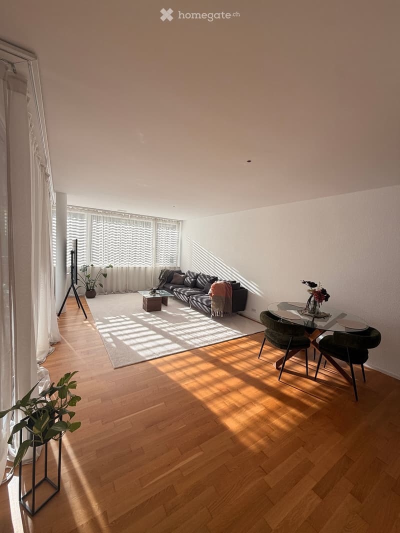 Bright living room with wooden flooring, large windows with white curtains, a black sectional sofa, a glass coffee table, a dining table with chairs, and plants.