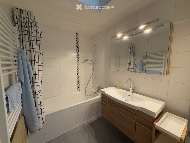 Modern bathroom with white tiles, a bathtub, a sink with a wooden cabinet, a large mirror with built-in lighting, and a towel on a radiator.