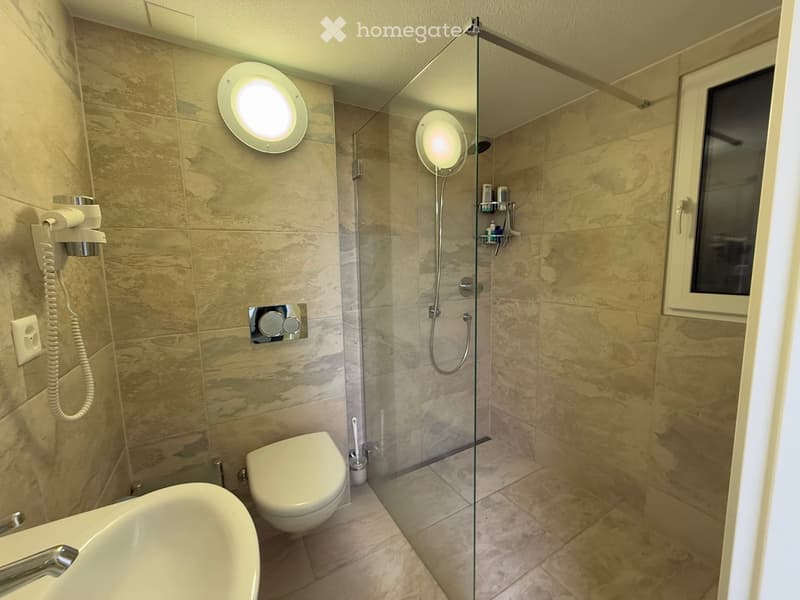 Modern bathroom with marble tiles, bathtub, toilet, glass-enclosed shower, and built-in shower shelf. Features two round wall-mounted lights and a hair dryer on the wall.