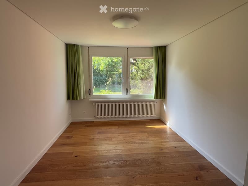 Empty room with wooden flooring, white walls, large windows with green curtains, and a radiator below the windows.