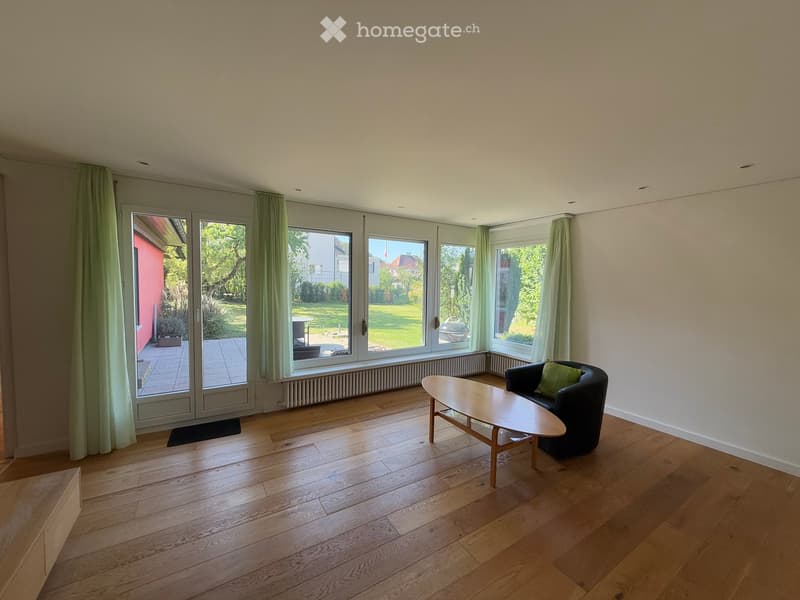 Bright living room with wooden flooring, large windows, and sliding glass doors leading to a garden. Features a black sofa with green cushions and a wooden coffee table.