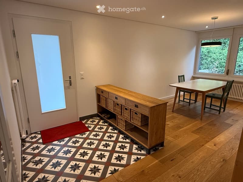 Bright room with wooden flooring, a large window, a dining table with chairs, a wooden cabinet with wicker baskets, and a patterned rug near the door.