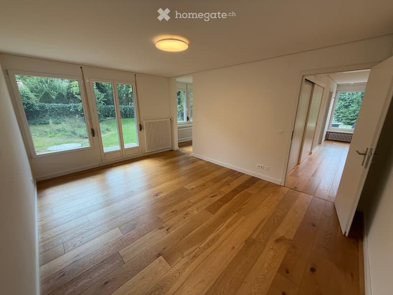 Empty room with wooden flooring, large windows, and a sliding door leading to a garden.