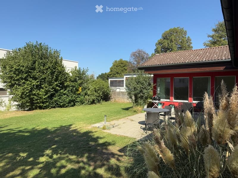 Well-maintained garden with a lawn, patio seating area, and a small red house with large windows in the background.