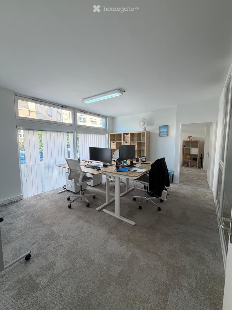 Bright, modern office space with large windows, a standing desk, two chairs, dual monitors, and shelves for storage.