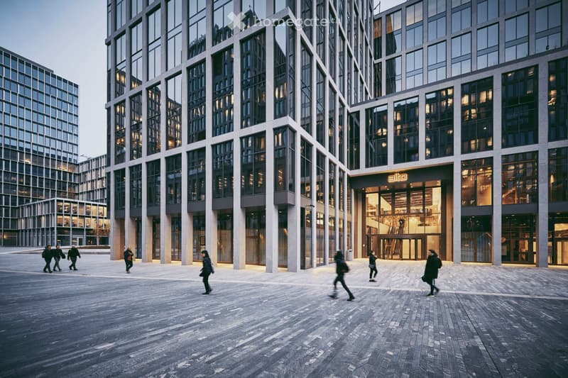 Modern multi-story building with large glass windows and a central entrance, surrounded by a paved plaza with people walking.
