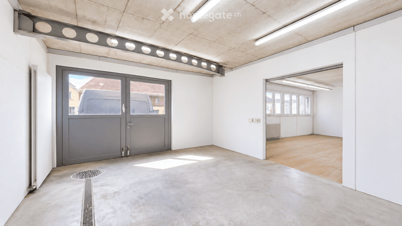 Empty garage with concrete floor, large grey door, and adjacent room with wooden flooring and white walls.
