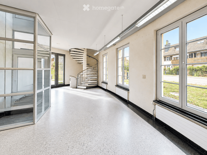 Modern, spacious hallway with large windows, a spiral staircase, and a glass elevator. The floor is carpeted, and the walls are light-colored.