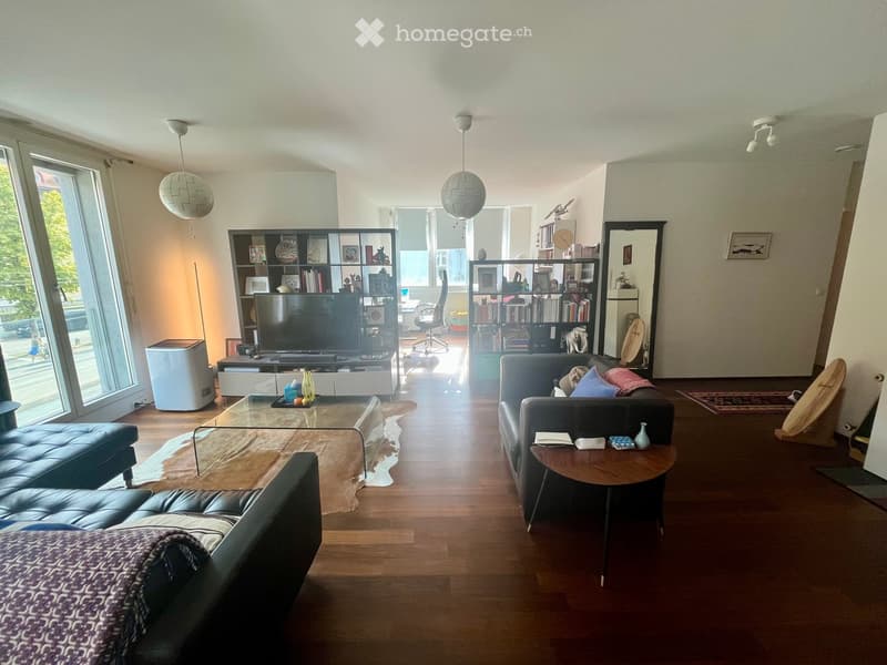 Modern living room with hardwood floors, large windows, a sofa, coffee table, bookshelf, and a television.