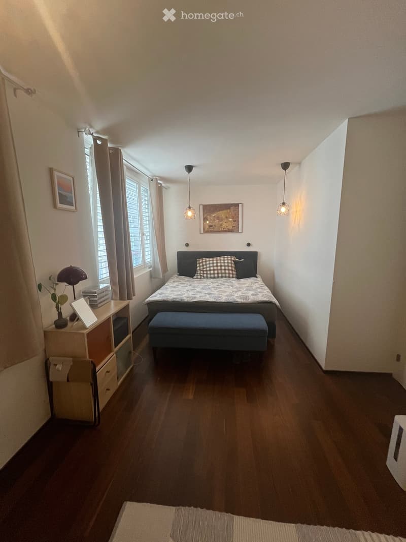 Modern bedroom with wooden flooring, large window with curtains, bed with dark headboard and patterned bedding, nightstand with lamp, stool, and decorative items.