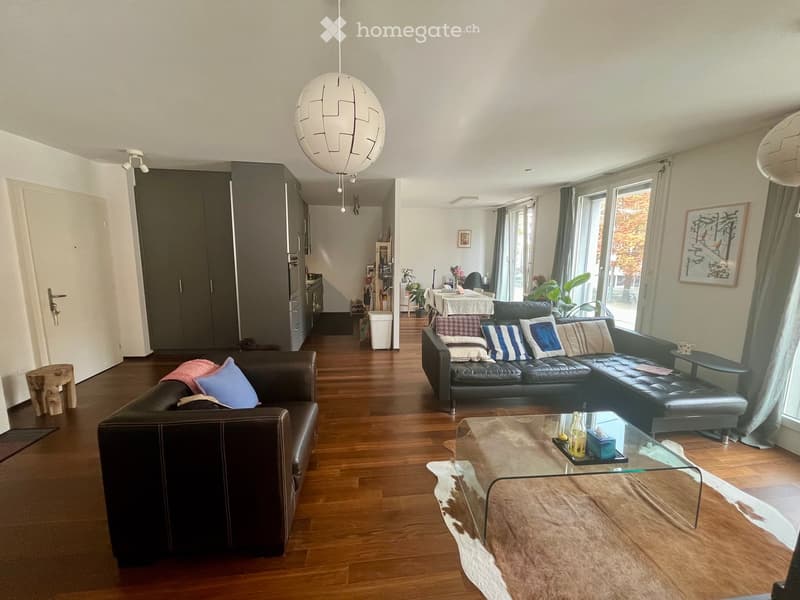 Modern living room with hardwood floors, leather sofas, glass coffee table, pendant lights, and large windows with curtains.