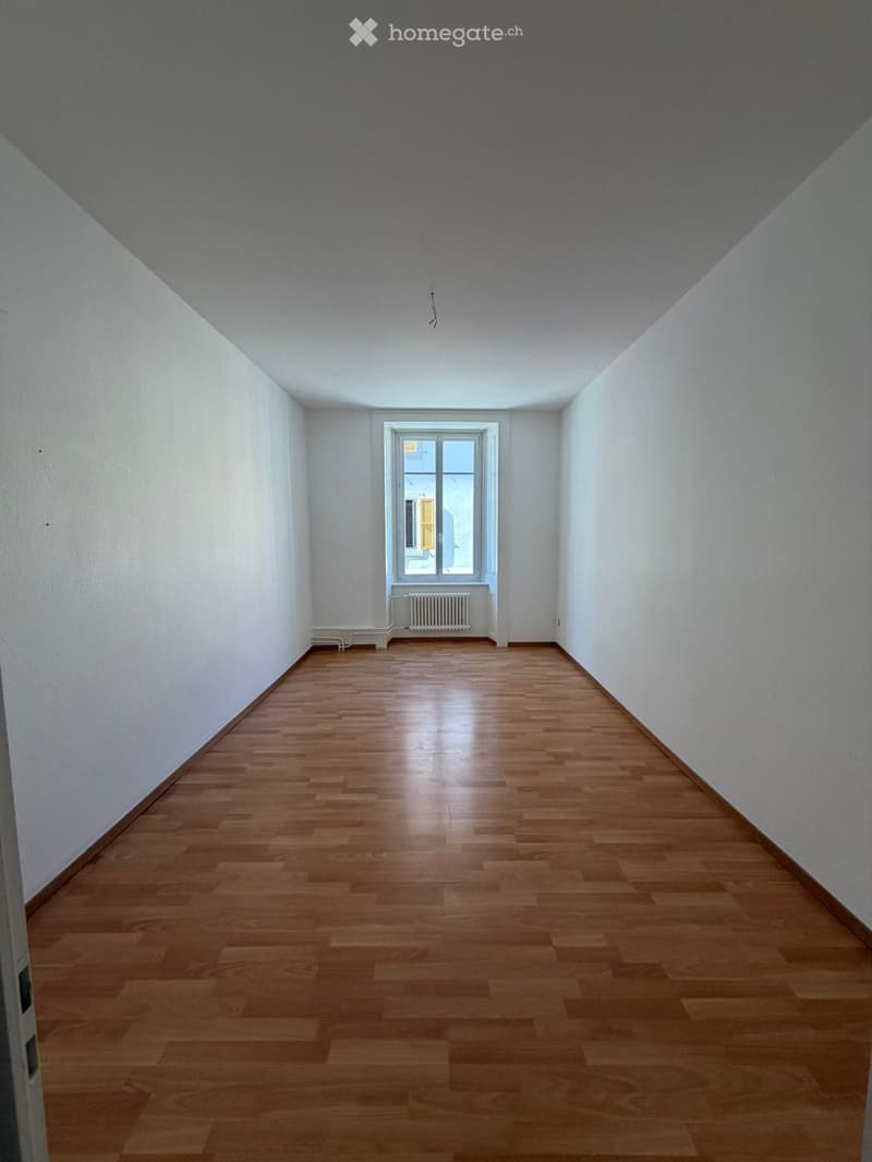 Empty room with wooden flooring, white walls, and a single window with a radiator below it.