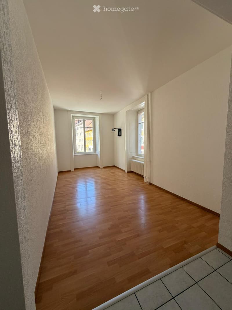 Empty room with wooden flooring, white walls, and two windows offering a view of the outside. There is a radiator under one window and a light fixture on the wall.