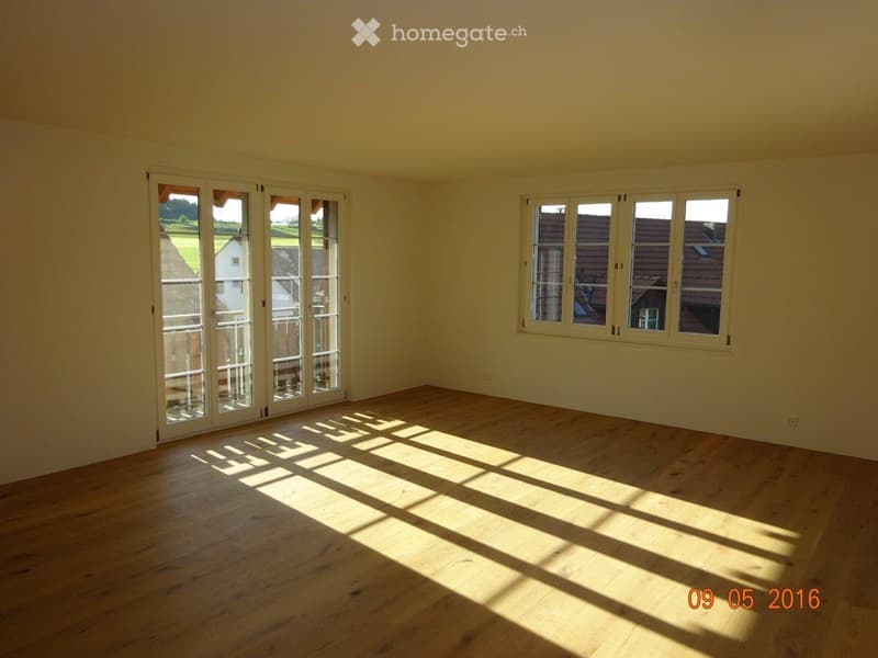 Empty room with wooden flooring and large windows, allowing natural light to fill the space. The room features a sliding glass door leading to an outdoor area.