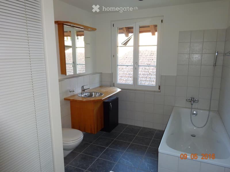 Bathroom with a bathtub, sink, toilet, and a mirror above the sink. The room has tiled walls and a window with a view of a tiled roof.
