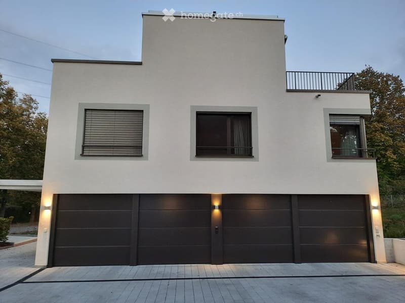 Modern two-story house with a flat roof, white facade, large garage doors, and a small balcony on the upper level.