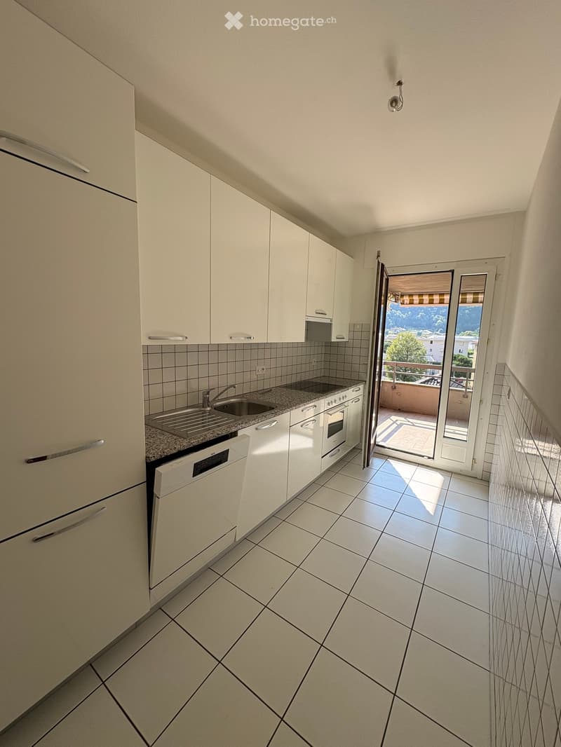 Modern kitchen with white cabinets, granite countertop, sink, oven, and dishwasher. Open sliding door leads to a balcony with outdoor view.
