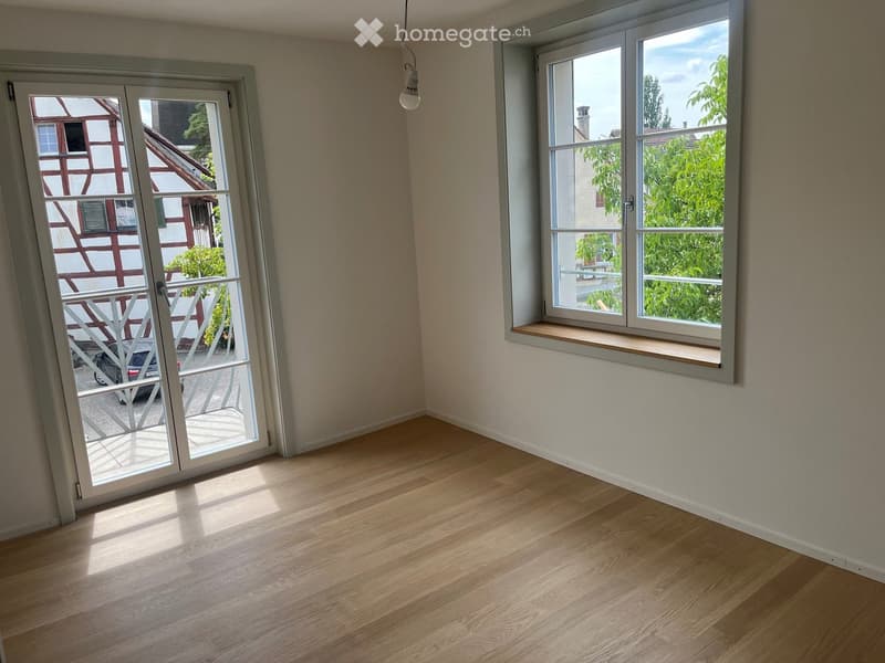 Empty room with wooden flooring, large windows, and a sliding glass door leading to a balcony.