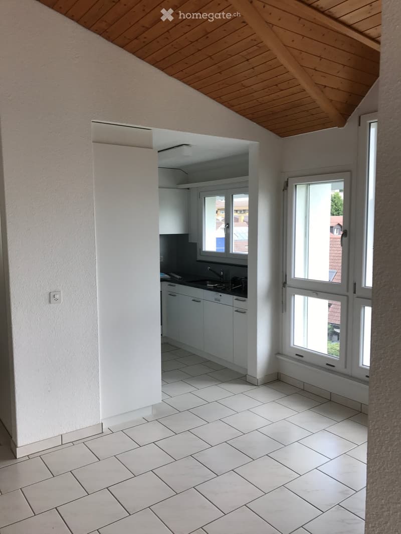 Empty room with white tiled floor, white walls, and a doorway leading to a small kitchen area with white cabinets and a sink.