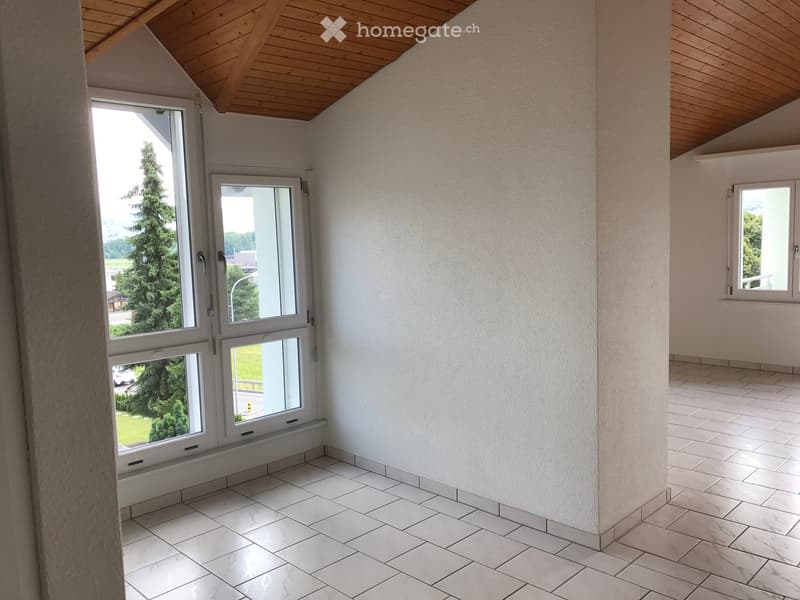 Empty room with white tiled flooring, white walls, and large windows offering a view of greenery outside. The ceiling has exposed wooden beams.