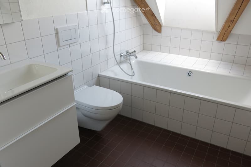 Modern bathroom with white tiled walls, a bathtub, toilet, and sink. The floor is covered with dark brown tiles, and the space features a window with wooden beams.