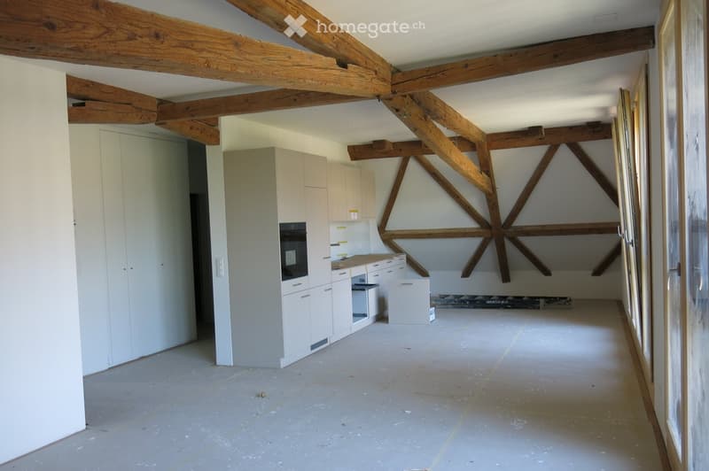 Empty room with exposed wooden beams, white cabinetry, and a partially constructed kitchen area. The floor is unfinished concrete, and the walls are painted white.