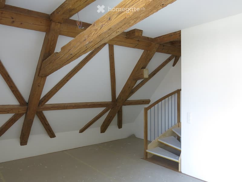 An attic space with exposed wooden beams and a small staircase leading up, featuring white walls and a concrete floor.
