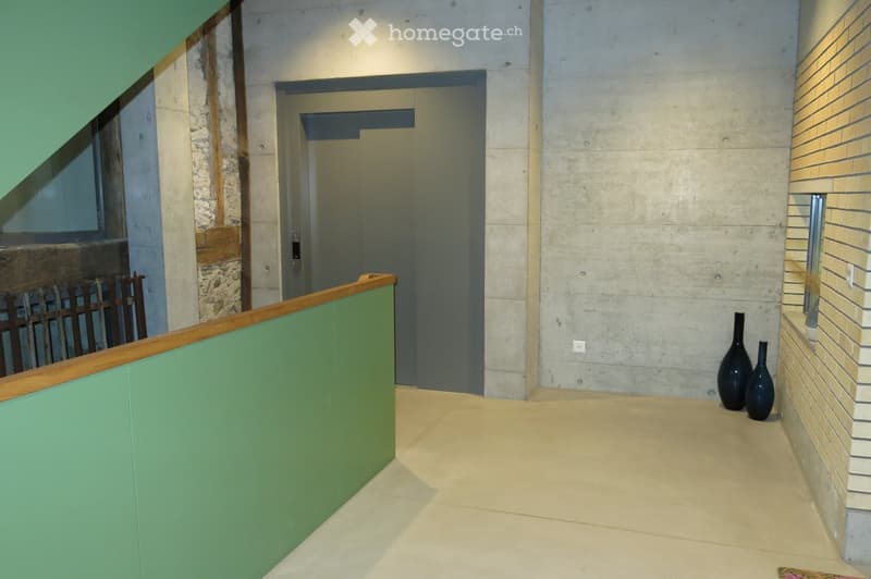 Modern hallway with concrete walls, tiled floor, green railing, and two black vases on a brick wall.