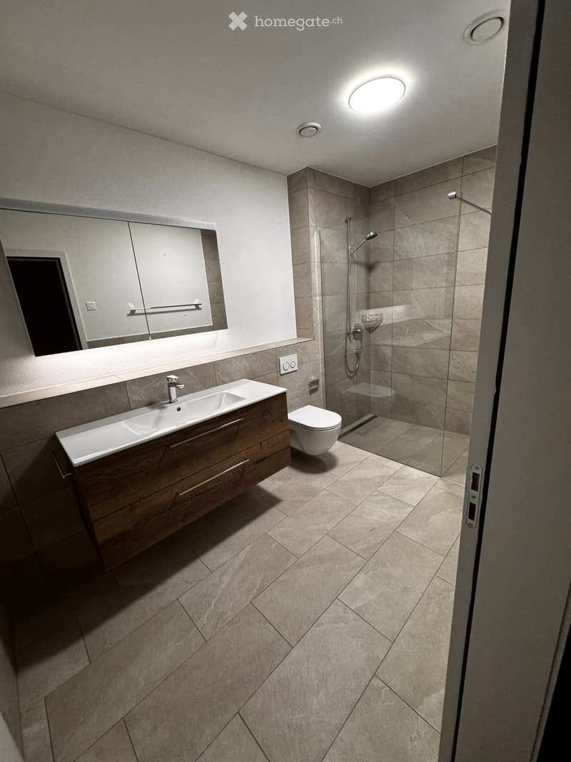 Modern bathroom with beige tiles, a wooden vanity with a sink, a toilet, and a glass-enclosed shower area.