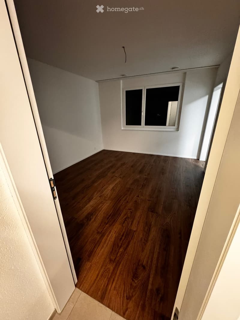 Empty room with wooden flooring, white walls, and a window with two panes. The room is unfurnished and has a door visible on the left side.