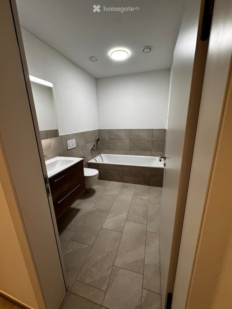 Modern bathroom with a bathtub, toilet, sink, and wooden cabinet. Features tiled walls and floor, recessed lighting, and a built-in shelf.
