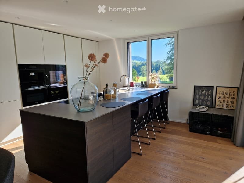 Modern kitchen with a dark wooden island, four stools, a glass vase with dried flowers, an oven, and a sink with a modern faucet. Large windows offer a scenic view.