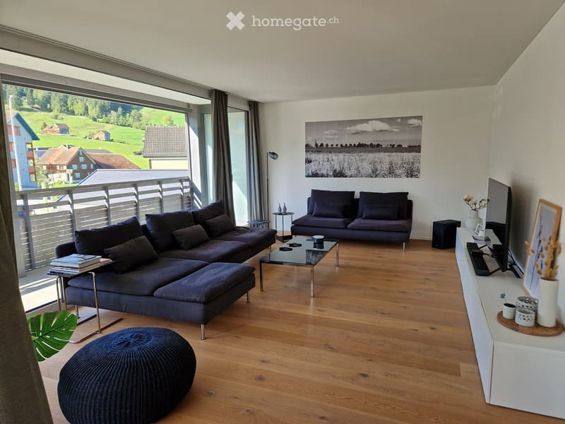 Modern living room with wooden flooring, large windows offering a scenic view, a dark blue sectional sofa, a coffee table, an ottoman, side tables, a floor lamp, and a television on a white console.