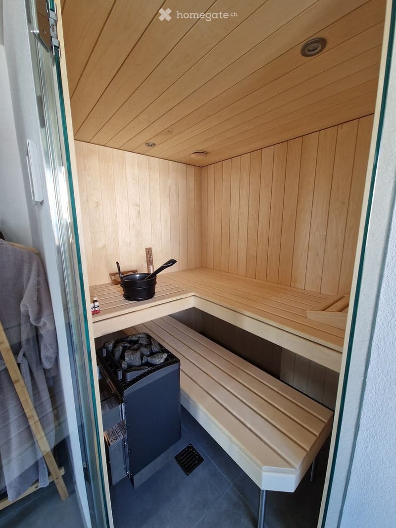 The image shows a small, wooden sauna with a wooden bench, wooden slats, a black pot on a wooden shelf, a wooden stove with rocks, and a wooden door. The sauna has a wooden ceiling and walls, and a wooden floor.