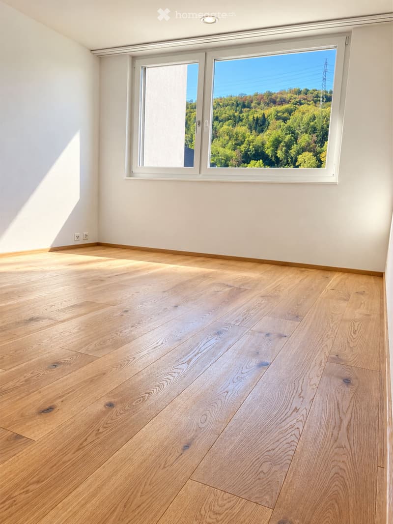 Empty room with light wooden flooring, white walls, and large windows offering a scenic view of greenery outside.