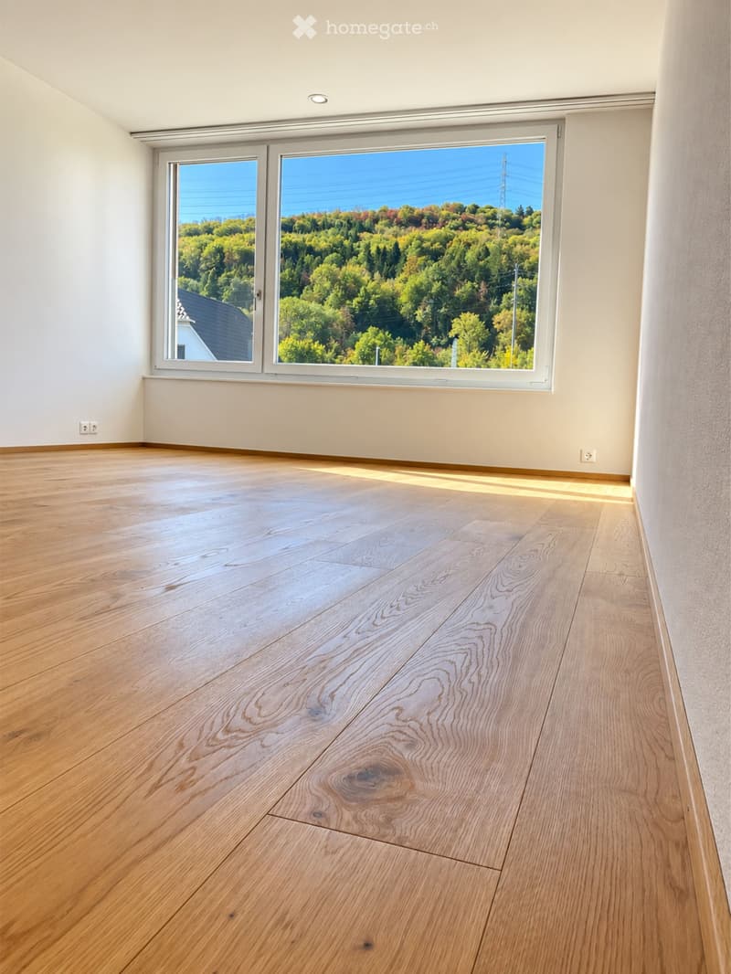 Empty room with light wooden flooring and large windows offering a scenic view of trees and a house.