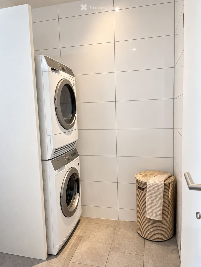 A small, modern laundry room with white tiled walls, a stacked washer and dryer, a woven basket with a towel, and a tiled floor.