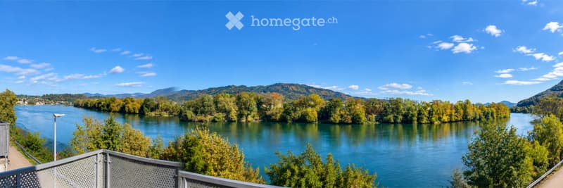 Balcony overlooking a river with surrounding trees and distant hills under a clear blue sky.